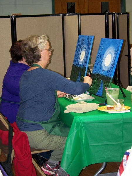 A painter in a green apron adding moonlight to a canvas of trees against a blue night sky