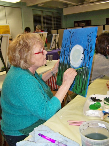A painter holding a canvas of a full moon through trees while adding grass in the foreground
